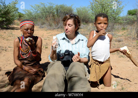 Turista femminile con Naro San donna e bambina mangiare dissetanti kombrua root. Kalahari Ghanzi regione, Botswana, Africa. La stagione secca, ottobre 2014. Foto Stock