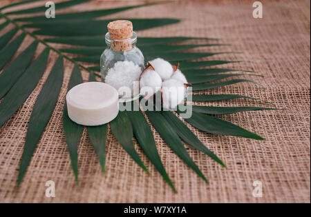 Sapone, sale marino in una bottiglia di vetro e cotone fiore su foglia di palma sfondo. Foto Stock