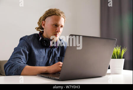 Bello studente con dreadlocks che indossano le cuffie seduti al tavolo del salotto, utilizzando il computer portatile a casa, guardando a schermo Foto Stock