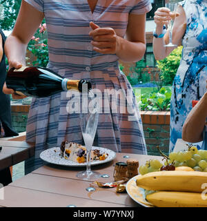 Una donna in un blu e bianca a strisce abito versa dello Champagne in un flute da champagne su un tavolo con dolci e frutta in un giardino esterno parte. Foto Stock