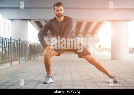 La vita è uno sport tutta la lunghezza di giovane e fiducioso uomo bearded in sportswear che allunga la sua gamba mentre sta in piedi all'aperto. Concetto di sport. Stile di vita sano e attivo Foto Stock