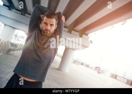 Riscaldamento prima dello scatto. Ritratto di giovane uomo bearded in sportswear che allunga il braccio mentre si alza all'aperto. Concetto di sport. Stile di vita sano e attivo Foto Stock