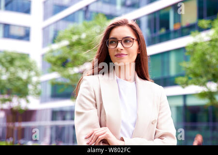 Colpo di bella giovane imprenditrice indossa blazer e occhiali rimanendo sulla strada con bracci ripiegati. Foto Stock
