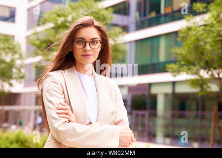 Colpo di bella giovane imprenditrice indossa blazer e occhiali rimanendo sulla strada con bracci ripiegati. Foto Stock