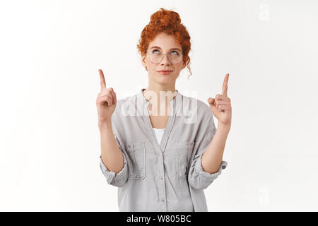 Curioso scaltri giovani redhead donna indossare gli occhiali, camicetta, cercando e rivolta verso l'alto interessati, smirk sly, avente interessanti piano, permanente backg bianco Foto Stock