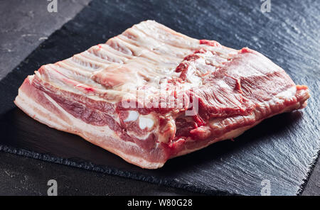 Carne di maiale cruda ventre con le costine e la pelle su un nero ardesia vassoio, vista da sopra, close-up Foto Stock