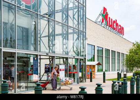 Ingresso al supermercato Auchan con logo e donna anziana che spinge il carrello completo, Budapest, Ungheria Foto Stock