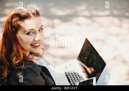 Close up ritratto di un bel giovane Taglie donna con capelli rossi lavora presso il suo portatile mentre è seduto nel parco guardando la telecamera a spalla Foto Stock