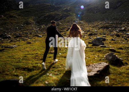 Irriconoscibile vista posteriore del Caucaso in sposa e lo sposo tenendo le mani e correndo in montagna contro sunrise. Foto Stock