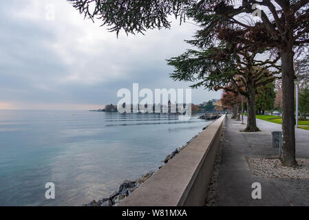 Tranquilla bella passerella naturale e la passeggiata lungo il lago sul lago di Ginevra e sullo sfondo di un nebbioso e nuvoloso e al crepuscolo cielo sopra acqua in inverno. Foto Stock