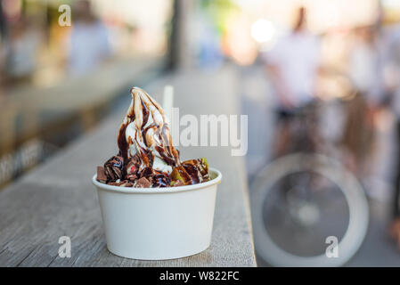 White Frozen Yogurt servire con topping di frutta e cioccolata in carta bianca cup, su legno tabella contatore su strada. Foto Stock