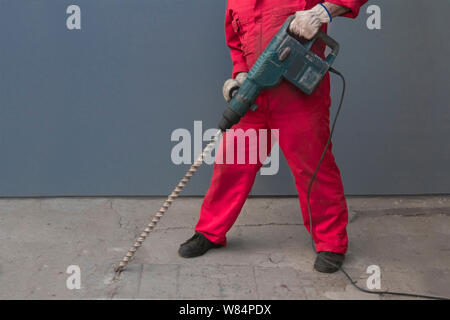 Un lavoratore in una tuta arancione lavora con un trapano con una punta lunga distrugge la pavimentazione in cemento. Un uomo tiene nelle sue mani un perforatore con un lungo b Foto Stock