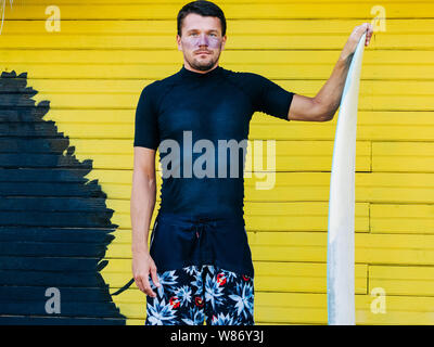 Ritratto di un surfista sulla faccia di un filtro solare, che indossa pantaloni corti e t-shirt nera in piedi con una tavola da surf. giallo sullo sfondo di legno. Foto Stock