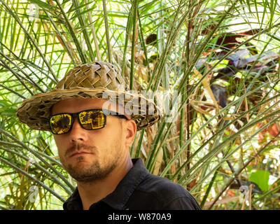 Closeup ritratto di un uomo che indossa un sombrero graffiato occhiali da sole in un verde giardino tropicale. Foto Stock