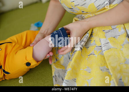 Giovani caucasici madre indossa un vintage abito giallo e aiutando a little baby boy ottenere pronto, con focus sulle mani il montaggio carino babbucce blu Foto Stock