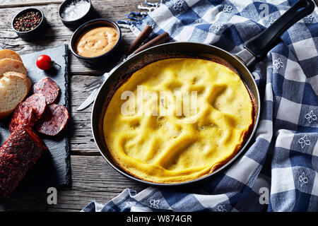 Frittata classica in una padella servita affettata con chorizo, pane e pomodori su un scheda di ardesia su un tavolo di legno con carta asciugatutto, vista da sopra, clo Foto Stock