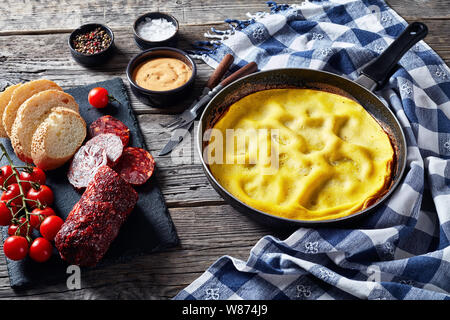 Frittata di caldo in una padella, affettato chorizo, pane e pomodori su un scheda di ardesia su un tavolo di legno con carta asciugatutto, vista da sopra, close-up Foto Stock