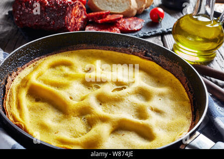Frittata di caldo in una padella, piccante a fette di carne di maiale salsiccia, il pane e i pomodori su una scheda di ardesia su un tavolo di legno, vista da sopra, close-up Foto Stock