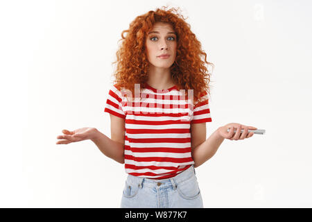 Clueless perplesso carino redhead elegante curly-ragazza dai capelli scrollare incerto non hanno idea di sollevamento lato dubbia tenere lo smartphone strano discutere messa Foto Stock