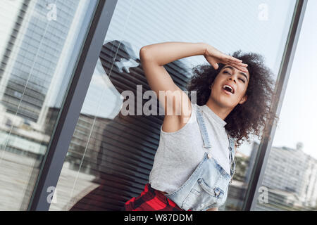 Giovane donna nella strada di città in piedi vicino alla finestra nasconde da Sun che cercano riposo aperto bocca riflessivo Foto Stock