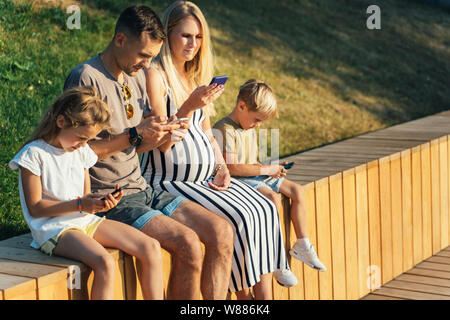 Immagine dei genitori con la figlia e il Figlio seduto sulla staccionata in legno con telefoni nelle mani Foto Stock