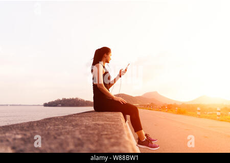 Vista laterale dello sport donna con gli auricolari di seduta sul muro dopo un pomeriggio eseguite. Un atleta femminile di prendere un periodo di riposo dopo un allenamento all'aperto Foto Stock