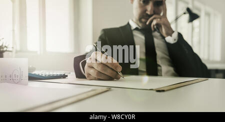 Preoccupato imprenditore seduto alla sua scrivania che lavora su imposte e documenti finanziari con la calcolatrice e le ricevute sulla sua scrivania. Foto Stock