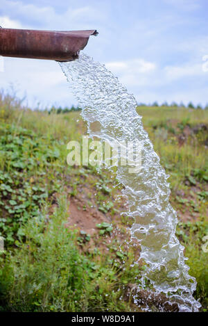 Un forte flusso di acqua pulita si riversa fuori un arrugginito vecchia tubazione nella campagna nel campo estivo Foto Stock