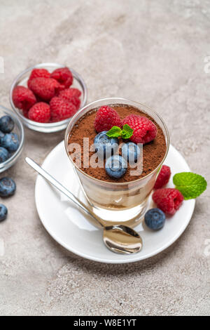 Tiramisù classico dessert con mirtilli e lamponi in un bicchiere e ciotole con frutti di bosco su sfondo di calcestruzzo Foto Stock