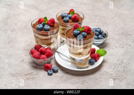 Tiramisù classico dessert con mirtilli e lamponi in un bicchiere e ciotole con frutti di bosco su sfondo di calcestruzzo Foto Stock