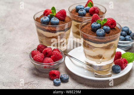 Tiramisù classico dessert con mirtilli e lamponi in un bicchiere e ciotole con frutti di bosco su sfondo di calcestruzzo Foto Stock