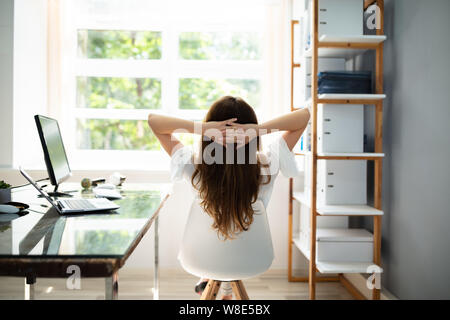 Vista posteriore di imprenditrice rilassato con le mani dietro la testa seduto in ufficio Foto Stock