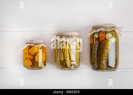 Salatura vari ortaggi in vasetti di vetro per lo stoccaggio a lungo termine. Conserve di verdure in vasi di vetro bianco su sfondo di legno. Vista dall'alto. Foto Stock