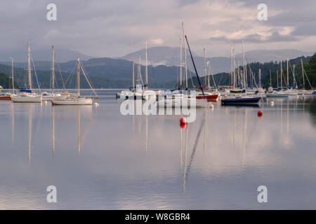 Barche a vela ormeggiata sulle calme acque di Windermere la mattina presto in agosto con il fells dietro Foto Stock