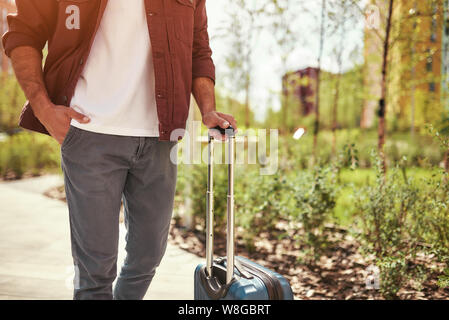 Andare all'aeroporto. Foto ritagliata di un uomo in abbigliamento casual tirando i suoi bagagli mentre a piedi attraverso la strada della citta'. Concetto di viaggio. Stile di vita Foto Stock