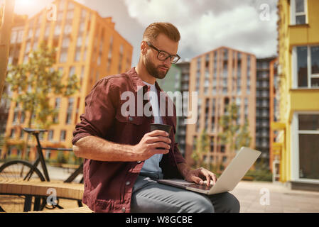 Ho bisogno di caffè. Bell'uomo con stoppia in abiti e occhiali casual che lavora su un computer portatile e che tiene una tazza di caffè mentre si siede sulla panca all'aperto. Stile di vita. Concetto di lavoro. Vita in città Foto Stock