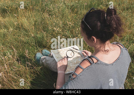 Ragazza esamina turistica attraverso una lente di ingrandimento qualcosa su una mappa con una penna in mano. Ci sono degli occhiali sulla testa. Il sole splende. Foto Stock