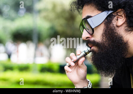 Uomo Barbuto fuma vape close up. Sigaretta elettronica concetto. Uomo con barba lunga e nuvole di fumo sembra rilassata. Uomo con barba e baffi sulla faccia di calma, rami su sfondo sfocato. Foto Stock