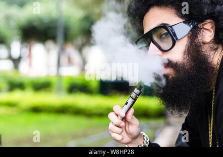 Uomo Barbuto fuma vape close up. Sigaretta elettronica concetto. Uomo con barba lunga e nuvole di fumo sembra rilassata. Uomo con barba e baffi sulla faccia di calma, rami su sfondo sfocato. Foto Stock