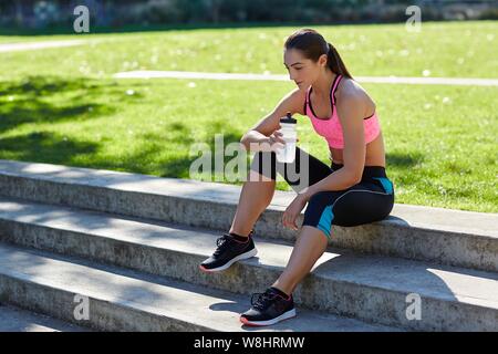 Giovane donna in abbigliamento sportivo in appoggio sui gradini con una bottiglia d'acqua. Foto Stock