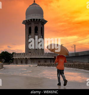 Turistico con un ombrello cammina verso vecchie di decenni di Clock Tower in un giorno di pioggia al mercato MoazzamJahi in Hyderabad, India Foto Stock