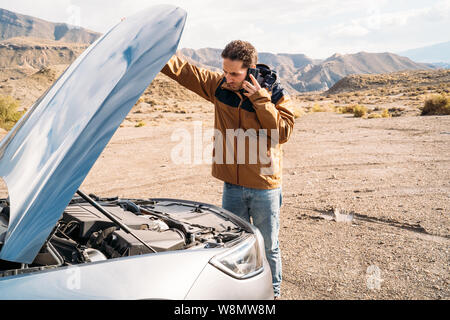 Un uomo con il cofano della sua auto aperto, parlando sul suo smart telefono cellulare nel mezzo del deserto - Concetto di vacanza Foto Stock