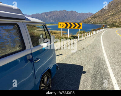 Un vintage blu e bianco camper parcheggiato nei pressi del bellissimo lago Hawea, con una curva e una velocità segnale di avvertimento in primo piano, contro un chiaro Foto Stock