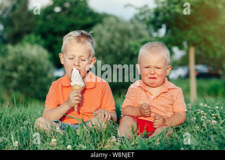 Due caucasica bambini divertenti ragazzi fratelli seduti insieme la condivisione di mangiare un gelato. Il Toddler più giovane pianto del bambino e il fratello più anziano teasing lui. L Foto Stock