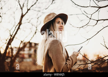 Vista laterale di affascinanti giovani donne in abito elegante e sorridente che guarda lontano mentre si utilizza lo smartphone e in piedi su sfondo sfocato di autunno coun Foto Stock