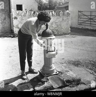 Degli anni Cinquanta, storico, per la stagione estiva e una signora camper riempimento di un travagliato bollitore in metallo con acqua proveniente da una pompa dell'acqua in un cantiere di fattoria, Inghilterra, Regno Unito. Foto Stock