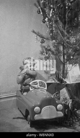 Degli anni Cinquanta, storico, un ragazzino abbraccia il suo orsacchiotti seduta nel suo giocattolo ride-su auto accanto a un albero di natale, Inghilterra, Regno Unito. Foto Stock