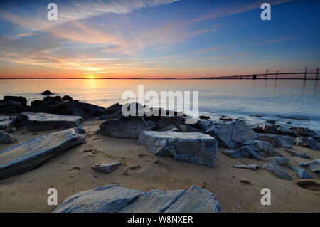 Alba sull'orizzonte a Chesapeake Bay Bridge in Maryland. Mattina colorate del cielo illuminano le rocce in primo piano lungo il litorale sabbioso. Foto Stock