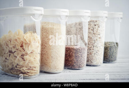 Vari cereali non cotte, cereali, fagioli e pasta per una cucina più sana in contenitori di plastica sul tavolo di legno, sfondo blu, close-up. Immagazzinaggio di prodotti alimentari c Foto Stock