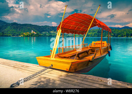 Fantastico legno tradizionale barca Pletna è ormeggiata presso il molo. Carino Pletna barca sul turchese del lago di Bled e la chiesa del pellegrinaggio con la piccola isola di ba Foto Stock